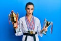 Beautiful brunette young woman wearing karate fighter uniform and medals holding trophy in shock face, looking skeptical and