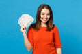 Beautiful brunette young woman wearing red orange dress holding fan of cash money in dollar banknotes isolated over blue