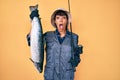 Beautiful brunettte fisher woman holding fishing rod and raw salmon afraid and shocked with surprise and amazed expression, fear Royalty Free Stock Photo