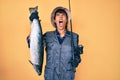 Beautiful brunettte fisher woman holding fishing rod and raw salmon angry and mad screaming frustrated and furious, shouting with Royalty Free Stock Photo