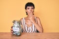Beautiful brunettte woman holding charity jar with money covering mouth with hand, shocked and afraid for mistake Royalty Free Stock Photo