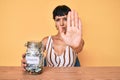 Beautiful brunettte woman holding charity jar with money with open hand doing stop sign with serious and confident expression,