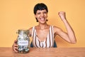 Beautiful brunettte woman holding charity jar with money screaming proud, celebrating victory and success very excited with raised Royalty Free Stock Photo