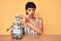 Beautiful brunettte woman holding charity jar with money in shock face, looking skeptical and sarcastic, surprised with open mouth Royalty Free Stock Photo