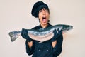 Beautiful brunettte woman professional chef holding fresh salmon fish angry and mad screaming frustrated and furious, shouting