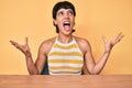 Beautiful brunettte woman sitting on the table over yellow background crazy and mad shouting and yelling with aggressive Royalty Free Stock Photo