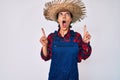 Beautiful brunettte woman wearing farmer clothes amazed and surprised looking up and pointing with fingers and raised arms Royalty Free Stock Photo