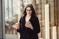 Beautiful business woman wearing glasses walking to the work with coffee and texting on phone outdoors. Royalty Free Stock Photo