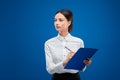 Beautiful businesswoman looking thoughtfully towards the left while writing on her blue clipboard,  on blue background Royalty Free Stock Photo