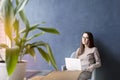 Beautiful businesswoman sitting in loft office using laptop on knees. Look and smile. Dark blue wall background, day light