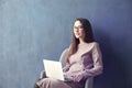 Beautiful businesswoman sitting in loft office using laptop on knees. Look and smile. Dark blue wall background, day light.