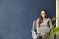 Beautiful businesswoman working on laptop while sitting in modern loft office. Dark blue wall background, day light.