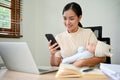 Beautiful busy Asian mother working from home, talking on the phone and holding her baby Royalty Free Stock Photo