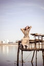 Beautiful caucasian blonde girl in light dress, sits on old wooden bridge over water against blue sky on city background Royalty Free Stock Photo