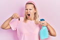 Beautiful caucasian blonde woman holding cockroach and pesticide sprayer angry and mad screaming frustrated and furious, shouting