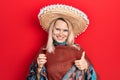 Beautiful caucasian blonde woman wearing festive mexican poncho drinking tequila shot smiling happy and positive, thumb up doing