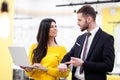 Beautiful Caucasian dark haired girl in yellow posing with businessman wearing suit. Happy colleagues with a laptop in a modern