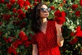 Beautiful caucasian girl with curly brunet hair in a trendy vintage red dress with polka dots with sunscreens stands near a rose Royalty Free Stock Photo