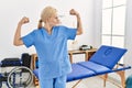 Beautiful caucasian physiotherapist woman working at pain recovery clinic showing arms muscles smiling proud Royalty Free Stock Photo