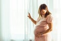 Beautiful caucasian pregnant woman touching her big belly in front of the window behind the curtain. Expectation of the childbirth