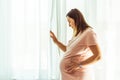 Beautiful caucasian pregnant woman touching her big belly in front of the window behind the curtain. Expectation of the childbirth Royalty Free Stock Photo
