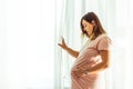 Beautiful caucasian pregnant woman touching her big belly in front of the window behind the curtain. Expectation of the childbirth