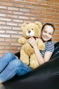 Beautiful caucasian teen girl sitting in black bean bag chair holding big brown teddy bear toy and smiling in camera. Casual outfi Royalty Free Stock Photo