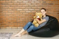 Beautiful caucasian teen girl sitting in black bean bag chair holding big brown teddy bear toy and smiling in camera. Casual outfi Royalty Free Stock Photo