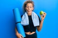 Beautiful caucasian teenager girl holding yoga mat and green apple making fish face with mouth and squinting eyes, crazy and Royalty Free Stock Photo