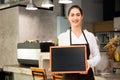 Beautiful Caucasian woman in barista apron holding empty blackboard sign inside coffee shop - ready to insert text