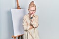 Beautiful caucasian woman with blond hair standing by painter easel stand thinking looking tired and bored with depression