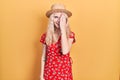 Beautiful caucasian woman with blond hair wearing summer hat covering one eye with hand, confident smile on face and surprise Royalty Free Stock Photo