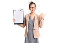 Beautiful caucasian woman with blonde hair holding clipboard with blank space pointing thumb up to the side smiling happy with Royalty Free Stock Photo