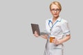 Beautiful Caucasian woman doctor or nurse holding a laptop pc computer and cup of coffee to go standing over grey Royalty Free Stock Photo