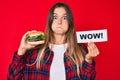 Beautiful caucasian woman eating a tasty classic burger holding wow text puffing cheeks with funny face