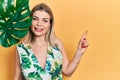 Beautiful caucasian woman with green plant leaf close to beautiful face smiling happy pointing with hand and finger to the side Royalty Free Stock Photo