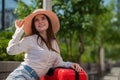 A beautiful Caucasian woman in a hat and shorts sits on a bench and holds a red suitcase and is waiting for a taxi or Royalty Free Stock Photo