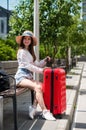 A beautiful Caucasian woman in a hat and shorts sits on a bench and holds a red suitcase and is waiting for a taxi or Royalty Free Stock Photo