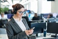 Beautiful caucasian woman in headset is holding a mobile phone while standing in open space office. Friendly female Royalty Free Stock Photo