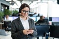 Beautiful caucasian woman in headset is holding a mobile phone while standing in open space office. Friendly female Royalty Free Stock Photo