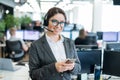 Beautiful caucasian woman in headset is holding a mobile phone while standing in open space office. Friendly female Royalty Free Stock Photo
