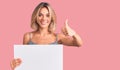 Beautiful caucasian woman holding blank empty banner smiling happy and positive, thumb up doing excellent and approval sign Royalty Free Stock Photo