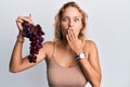 Beautiful caucasian woman holding branch of fresh grapes covering mouth with hand, shocked and afraid for mistake Royalty Free Stock Photo