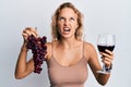 Beautiful caucasian woman holding branch of fresh grapes and red wine angry and mad screaming frustrated and furious, shouting Royalty Free Stock Photo