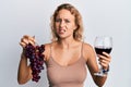 Beautiful caucasian woman holding branch of fresh grapes and red wine clueless and confused expression Royalty Free Stock Photo