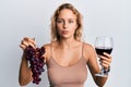 Beautiful caucasian woman holding branch of fresh grapes and red wine relaxed with serious expression on face Royalty Free Stock Photo