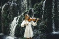 Beautiful Caucasian woman playing violin near waterfall. Music and art concept. Charming female with long blond hair wearing white Royalty Free Stock Photo