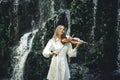 Beautiful Caucasian woman playing violin near waterfall. Music and art concept. Charming female wearing white dress in nature. Royalty Free Stock Photo