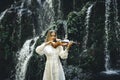 Beautiful Caucasian woman playing violin near waterfall. Music and art concept. Female wearing white dress in nature. Water splash Royalty Free Stock Photo