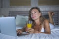 Beautiful Caucasian woman 30s lying on bed at night in home bedroom using internet at laptop computer drinking healthy orange juic Royalty Free Stock Photo